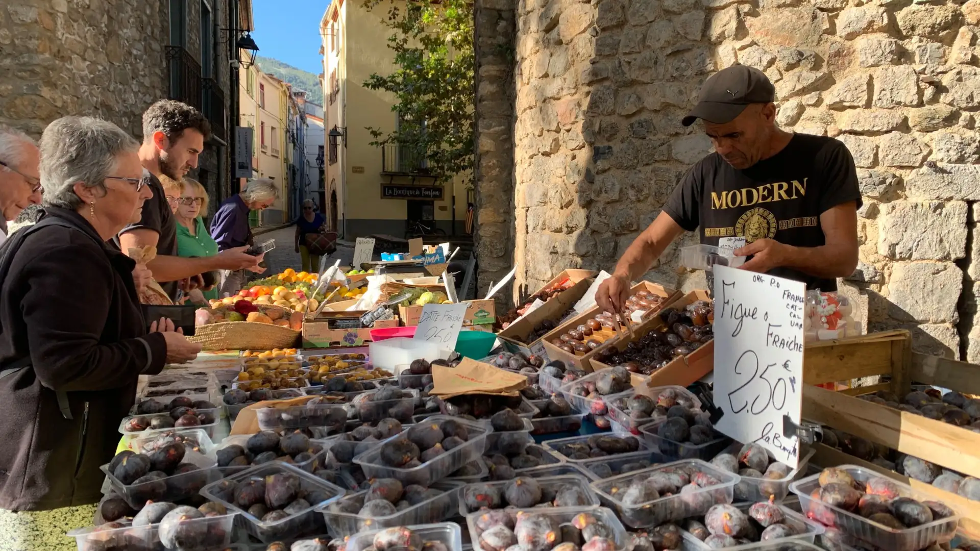 Marché de village animé sous une arche en pierre, étals de fruits et habitants